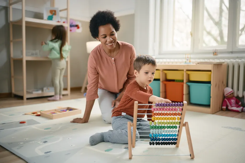 A toddler slides beads along a wooden abacus on a play mat while a smiling practitioner kneels beside him