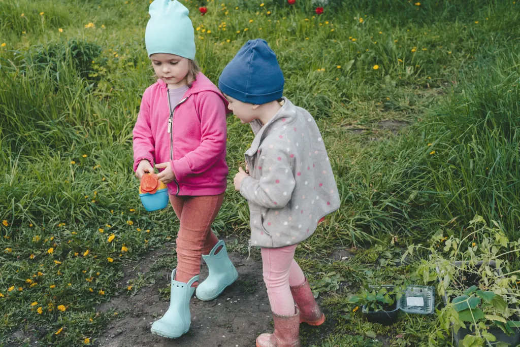 Two children in hats, jackets and wellies carry a watering can across the garden to the seedlings