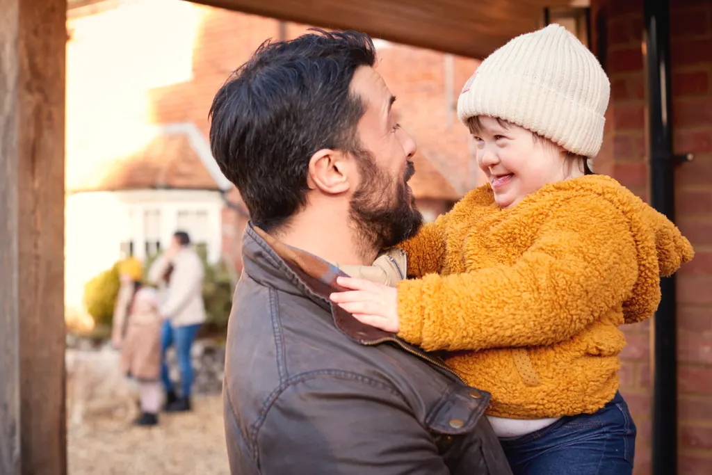 A smiling father holds up his toddler in a woolly hat and fleece coat under a brick porch at pick up